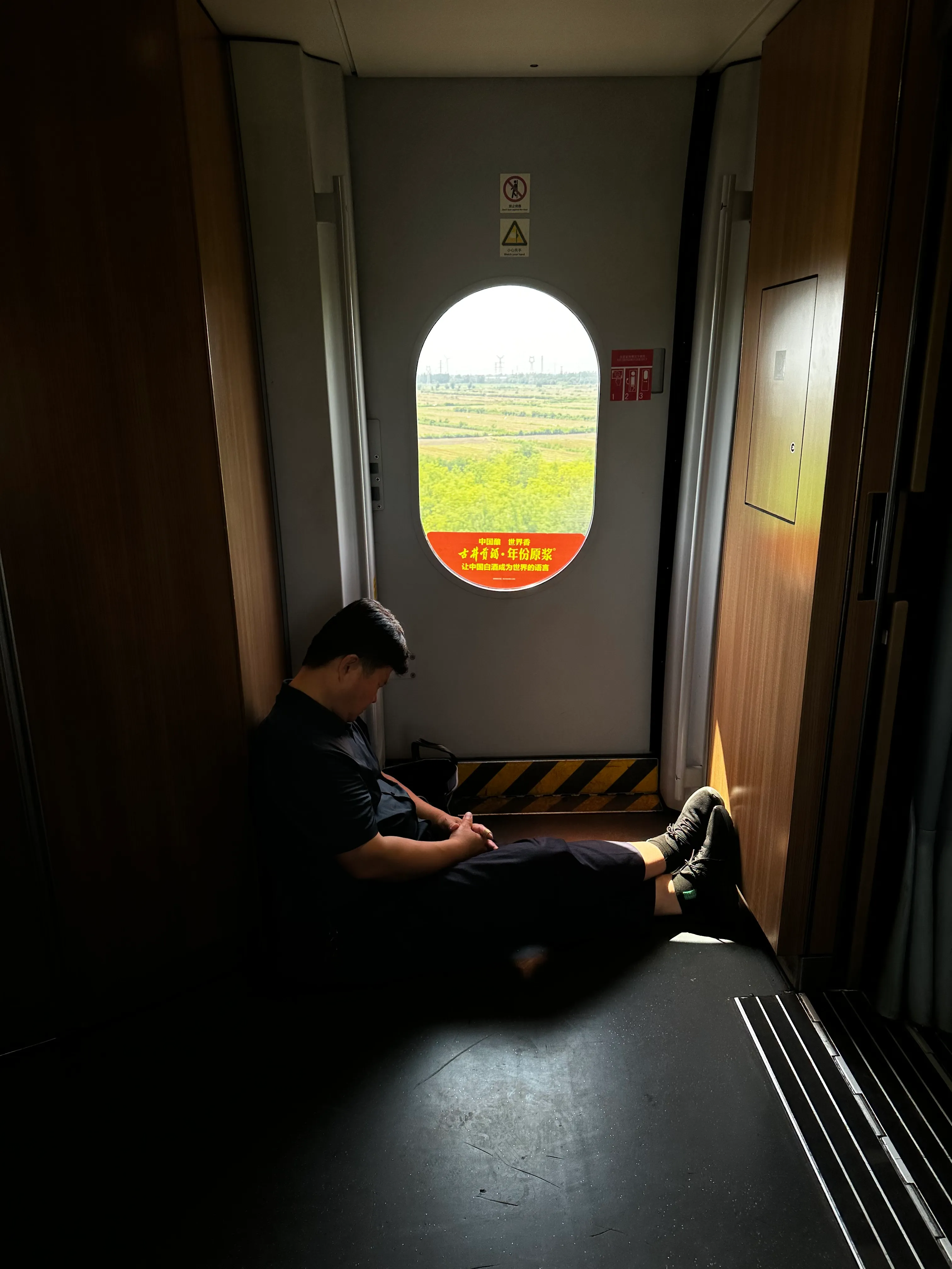 列车连接处，一位乘客在窗边休息，窗外是田野